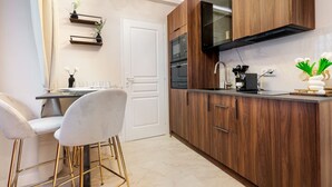 Een moderne keuken met houten kastjes, een zwarte magnetron en een roestvrijstalen spoelbak. Er staan twee witte stoelen met gouden poten bij de aanrecht, waar ook een klein plantje staat.