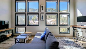 Ein modernes Wohnzimmer mit einer großen Couch, einem Fernseher und mehreren Fenstern mit Blick auf eine bewaldete Landschaft.