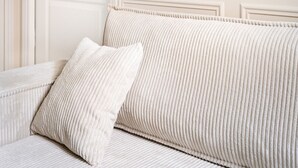 Un canapé blanc recouvert d’un tissu texturé, accompagné d’un coussin assorti, et un luminaire doré fixé au mur, placé au-dessus d’une porte blanche dotée d’une poignée ronde.