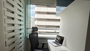 A modern office space with a white desk, black office chair, and a laptop. There is a window with blinds, and a view of a building outside. - Get a Flat - 604  Ibirapuera (São Paulo)
