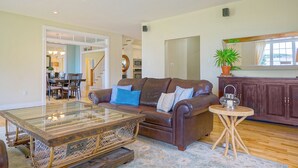 A spacious living room with a leather sofa, a glass coffee table, and a wooden side table. A dining area with a chandelier is visible in the background. - Harbourview Retreat - Waterview Luxury Home Steps from the Inn at Bay Fortune (Rock Barra)