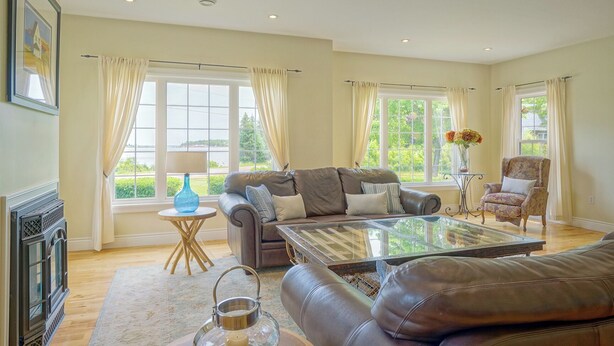 A spacious living room with a leather sofa, a glass coffee table, and a fireplace. There are large windows with curtains, a side table with a lamp, and a chair. - Harbourview Retreat - Waterview Luxury Home Steps from the Inn at Bay Fortune (Rock Barra)