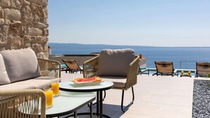 Una terraza con un muro de piedra, una mesa redonda con un plato de comida y un vaso de zumo de naranja, y varias sillas de mimbre.