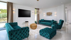 Una sala de estar moderna con un televisor de pantalla plana grande, muebles tapizados en color verde azulado, una araña y vistas al exterior con vegetación.