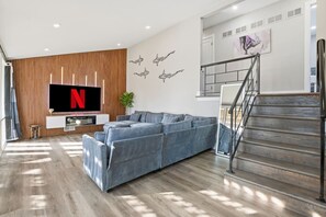Ein modernes Wohnzimmer mit einer großen Eckcouch, einem flachbildfernseher an einer Holzwand, einer Treppe mit Metallgeländer und einer minimalistischen Einrichtung.