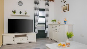 A living room with a flat-screen TV, a white cabinet, a small shelf with plants, a door with curtains, and a framed picture on the wall. - Holiday home Rozara by Villas Guide (Primošten)