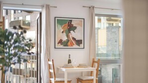 A dining area with a table set for two, a framed abstract painting on the wall, and a view of buildings through the window. - Bywell Apartment, Canning Town, London (Greater London)