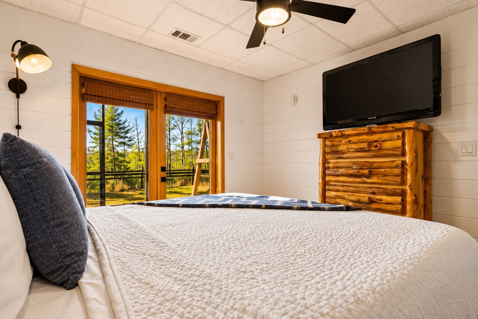 Een slaapkamer met een bed, een aan de muur bevestigde televisie, een plafondventilator en een raam met uitzicht op bomen.