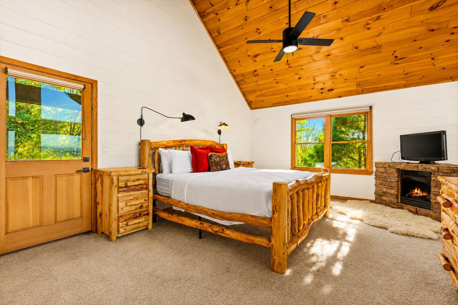 A bedroom with a wooden bed, a television, a fireplace, and large windows.