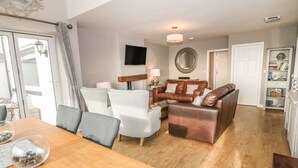Uma sala de estar moderna com uma mesa de jantar de madeira, cadeiras claras, um sofá de couro marrom e uma televisão de tela plana fixada na parede.