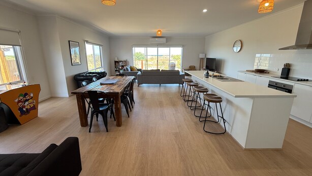 A spacious living area with a large wooden dining table, a kitchen with a white countertop, and a view of the outdoors through large windows.