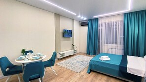 A modern hotel room with a bed, a dining table with chairs, a TV mounted on the wall, and a large window with curtains. - Cozy 1-bedroom apartment in a fantastic Almaty neighborhood (Almaty)