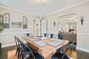 Un'area pranzo con tavolo in legno, sedie nere e un lampadario. Si vedono quadri appesi al muro e una pianta a sinistra.