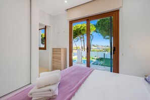 Un dormitorio con cama, cómoda de madera y vistas a una piscina y árboles a través de una puerta corrediza de vidrio.