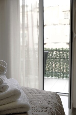 Ein ordentlich gefaltetes weißes Handtuch auf einem Bett mit einer gesteppten Decke, ein Fenster mit transparenten Vorhängen und Blick auf ein Gebäude und Grünfläche im Freien.