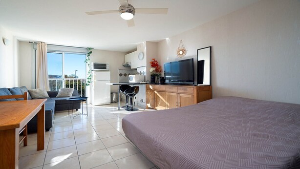 A modern hotel room with a large bed, a dining area with a table and chairs, a kitchenette, a TV, and a balcony with a view. - Cute little studio for 4 people, parking (Agde)