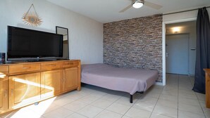A bedroom with a bed, a wooden dresser, a flat-screen TV, a stone wall, and a ceiling fan. - Cute little studio for 4 people, parking (Agde)