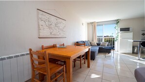 A dining area with a wooden table and chairs, a sofa with cushions, a map on the wall, and a kitchen with appliances. - Cute little studio for 4 people, parking (Agde)