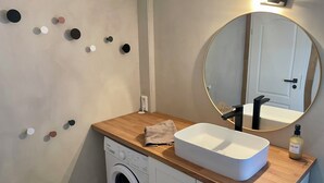 Een moderne badkamer met een houten aanrecht, een witte wasbak, een ronde spiegel, een wasmachine en een zeepdispenser aan de muur.