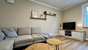 Een woonkamer met een grijze bank, twee ronde houten tafels, een televisie op een witte kast, en een raam met zonwering.