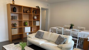 A living room with a white leather sofa, a marble coffee table, a wooden bookshelf, and a dining area with a white table and chairs. - Border Lodge - Comfort just a stone's throw from the border (Évrange)