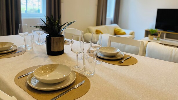 A dining table set with plates, glasses, and cutlery, a potted plant, and a sofa in the background. - Border Lodge - Comfort just a stone's throw from the border (Évrange)
