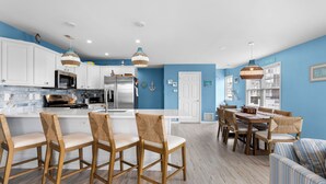 Een moderne keuken met witte kasten, een centraal eiland met barstoelen, en een eethoek met houten stoelen en een gestreept bankje.