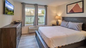 Un dormitorio con cama, cómoda, ventana con cortinas, televisor montado en la pared y un cuadro.