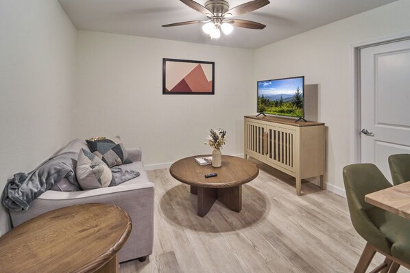 A modern living room with a ceiling fan, a television on a wooden cabinet, a round coffee table, a sofa with cushions, and a dining table with chairs. - Ruby 106: 1BD | Great Location+ Shared Amenities! (Pigeon Forge)