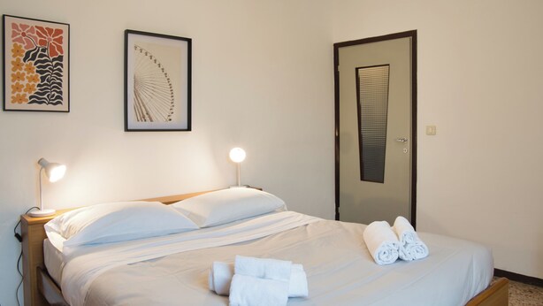 A neatly made bed with white linens and rolled towels, two framed artworks on the wall, and a door with a window. - Barzio - Garden - Primula (Barzio)