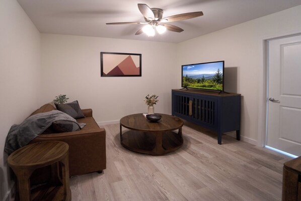 Un salon avec un canapé marron, une table basse ronde en bois, un téléviseur posé sur un meuble et un ventilateur de plafond.