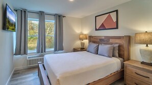 A bedroom with a wooden bed, a nightstand, a wall-mounted TV, and a window with curtains. - Ruby 109: 1BD | Great Location + Shared Amenities! (Pigeon Forge)