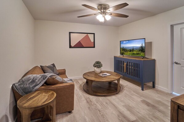 A living room with a brown sofa, a coffee table, a television on a blue cabinet, and a ceiling fan. - Ruby 112: 1BD | Great Location + Shared Amenities! (Pigeon Forge)