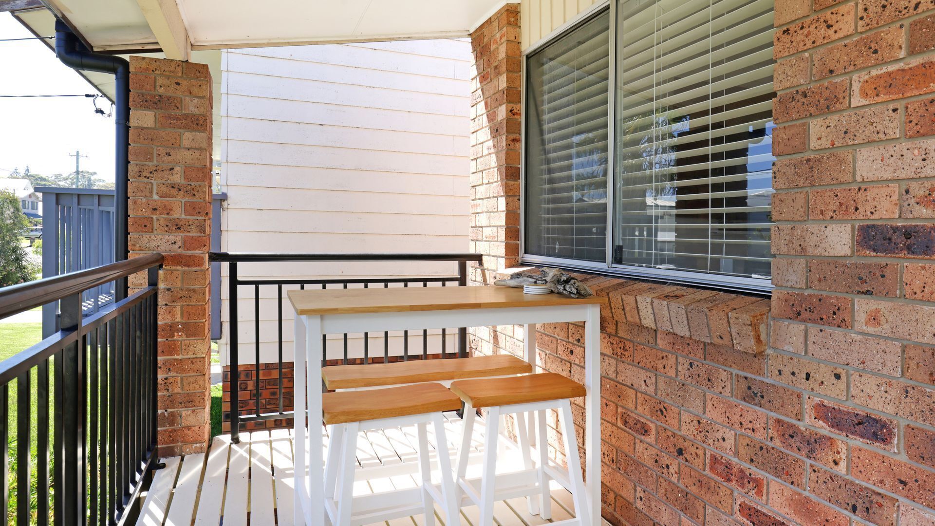 A balcony with a wooden table and two chairs, a window with blinds, and a brick wall.