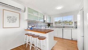 Cozinha moderna com armários brancos, bancada de madeira e banquetas. Há uma janela com vista para árvores e uma parede branca com uma citação emoldurada.