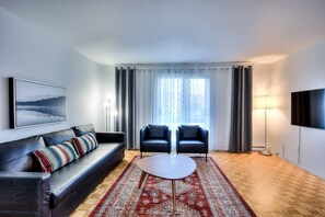 A modern living room with a sofa, two armchairs, a coffee table, a large window with curtains, a flat-screen TV, and a painting on the wall.