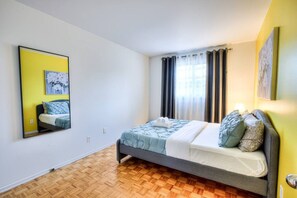 A bedroom with a bed, a mirror, a window with curtains, and a wooden floor.