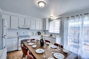 A modern kitchen with white cabinets, a wooden dining table set for four, and a large window with curtains.