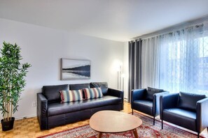 Una sala de estar moderna con un sofá de cuero negro, una mesa redonda y dos sillones. Un ventanal con cortinas traslúcidas deja pasar la luz natural.