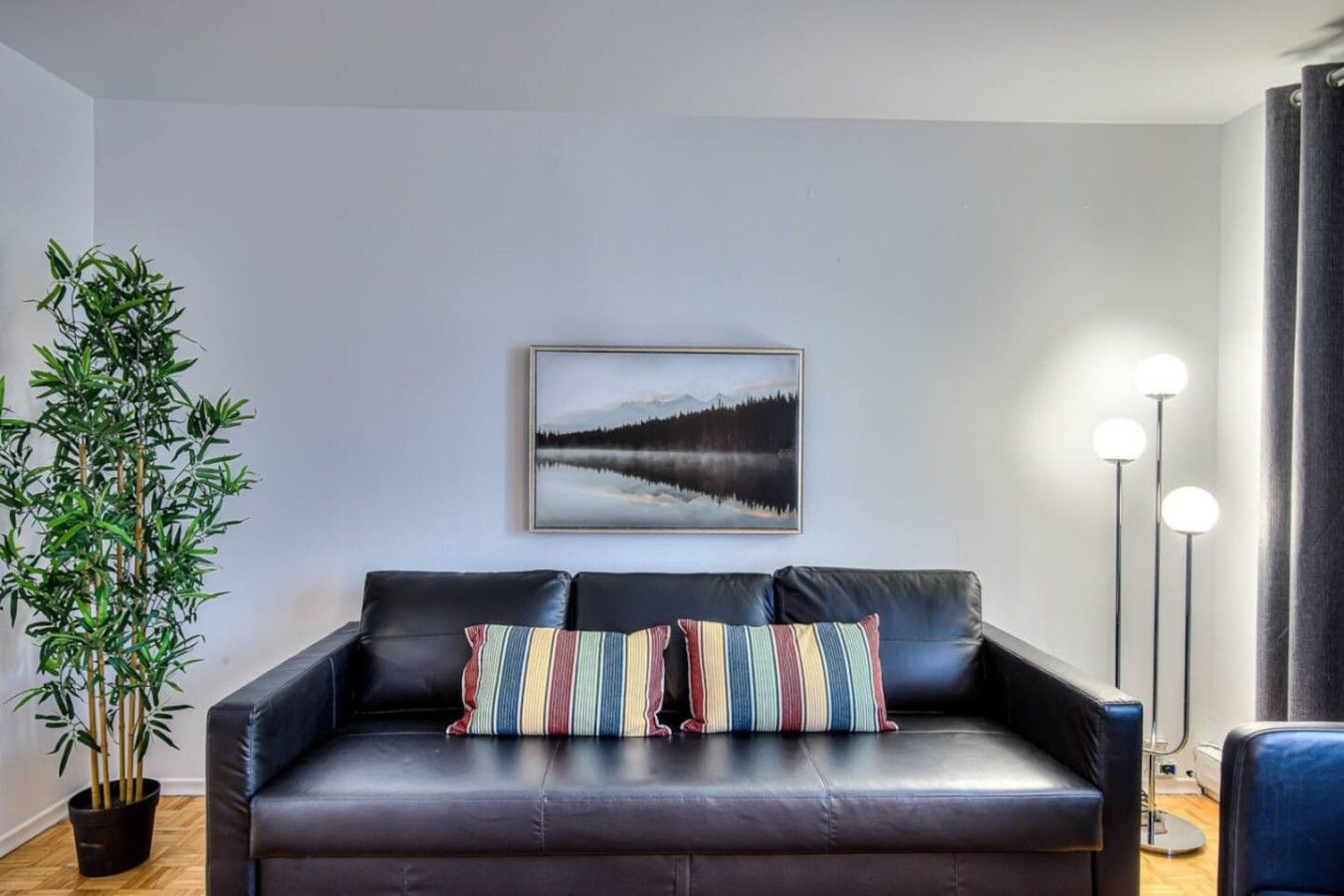 A modern living room with a black leather sofa, a framed landscape painting, a tall potted plant, and a floor lamp.