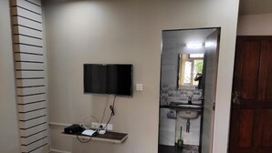 Ein Schlafzimmer mit Bett, einem an der Wand montierten Fernseher, einer kleinen Ablagefläche mit elektronischen Geräten und einem durch eine Glastür einsehbaren Badezimmer.