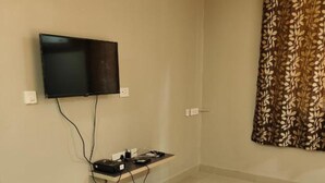 Ein Hotelzimmer mit einem an der Wand befestigten Flachbildfernseher, einer kleinen Ablagefläche mit elektronischen Geräten und einem gemusterten Vorhang.
