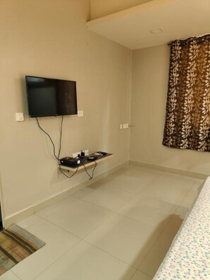 A hotel room with a flat-screen TV mounted on the wall, a small shelf with electronic devices, and a patterned curtain. - Deluxe Double Room in marvelous Colva with AC (Colva)