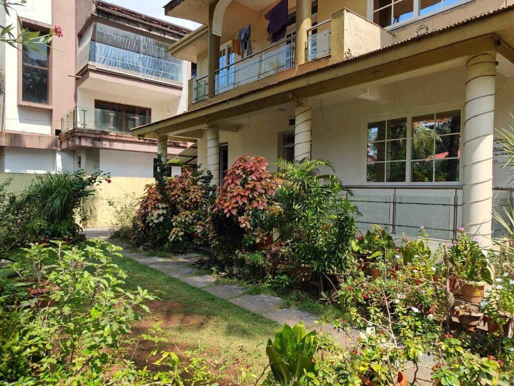 A two-story building with a covered balcony, a garden with various plants, and a pathway leading to the entrance.