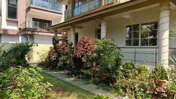 A two-story building with a covered balcony, a garden with various plants, and a pathway leading to the entrance.