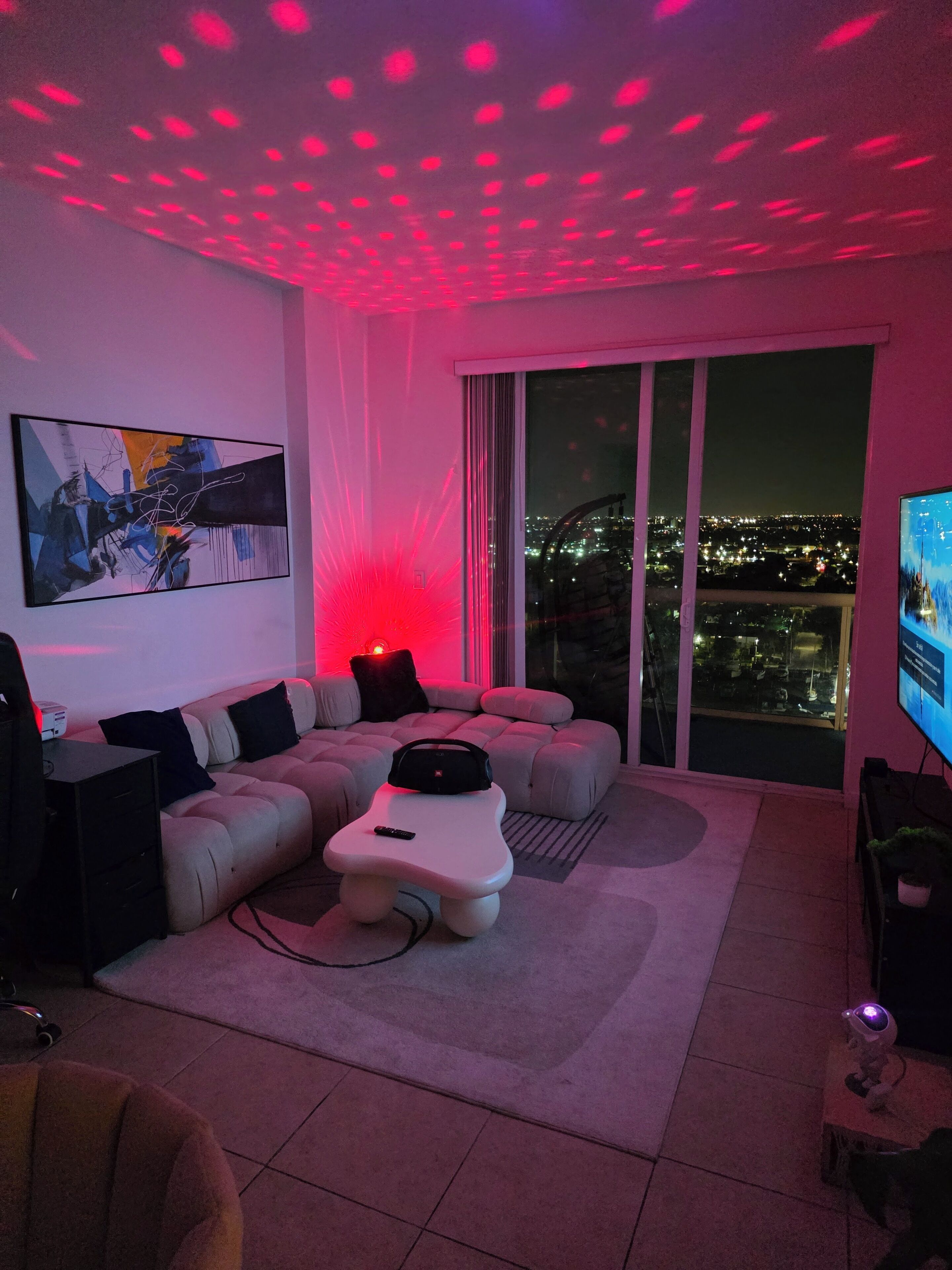 Una sala de estar moderna con un sofá modular, una mesa de centro blanca y un amplio ventanal con vistas a un paisaje urbano nocturno.