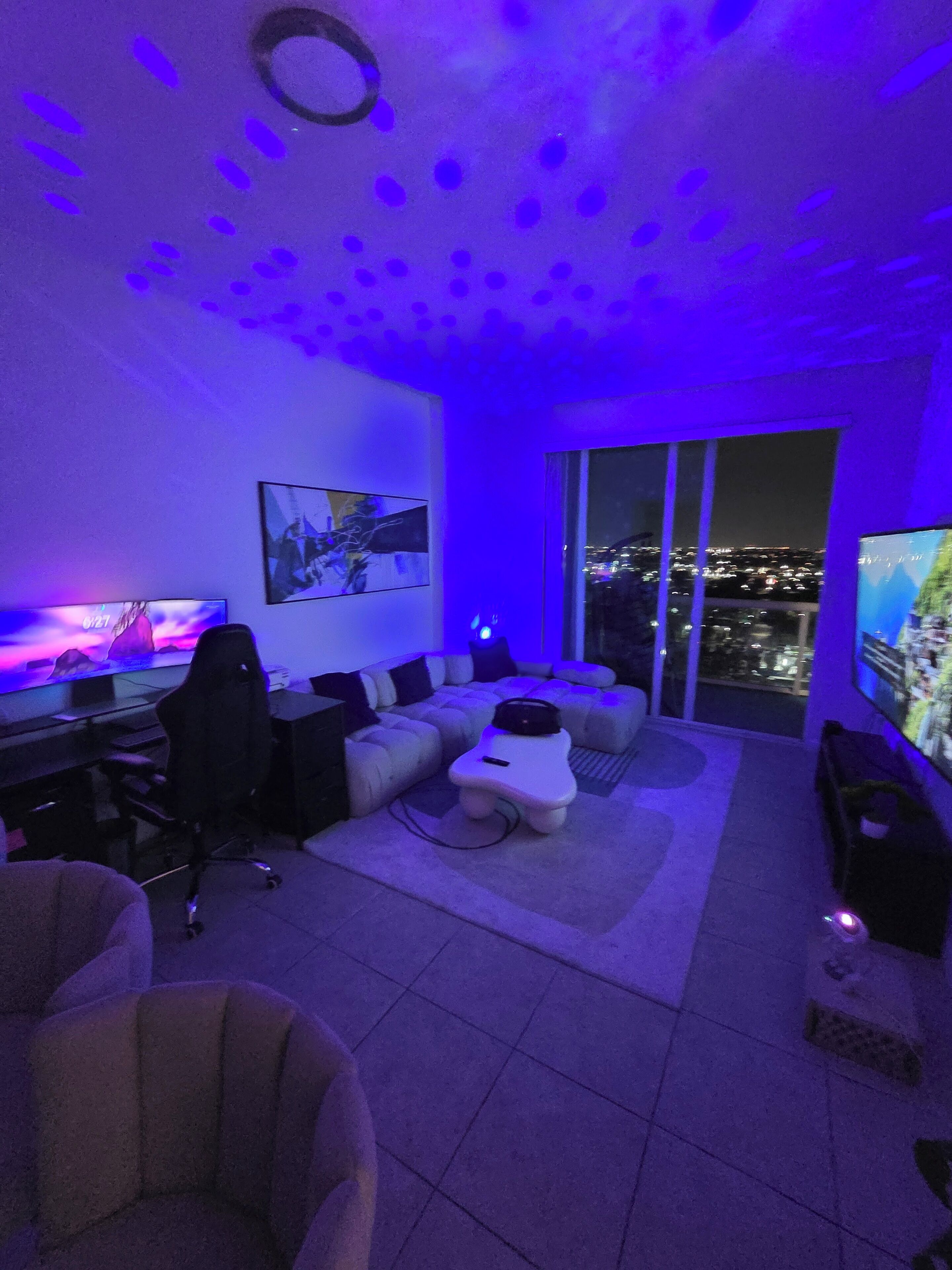 Ein modernes Wohnzimmer mit großem Flachbildfernseher, einer Couch, einem Couchtisch und einem Gaming-Sessel. Durch die großen Fenster ist die Stadt zu sehen.
