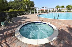 Ein Whirlpool, umgeben von einer Ziegelterrasse, mit einem Pool und Liegestühlen im Hintergrund.
