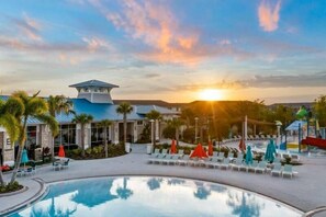 Zona de piscina de un resort con sillones de descanso y sombrillas, un edificio con techo azul y un atardecer de fondo.