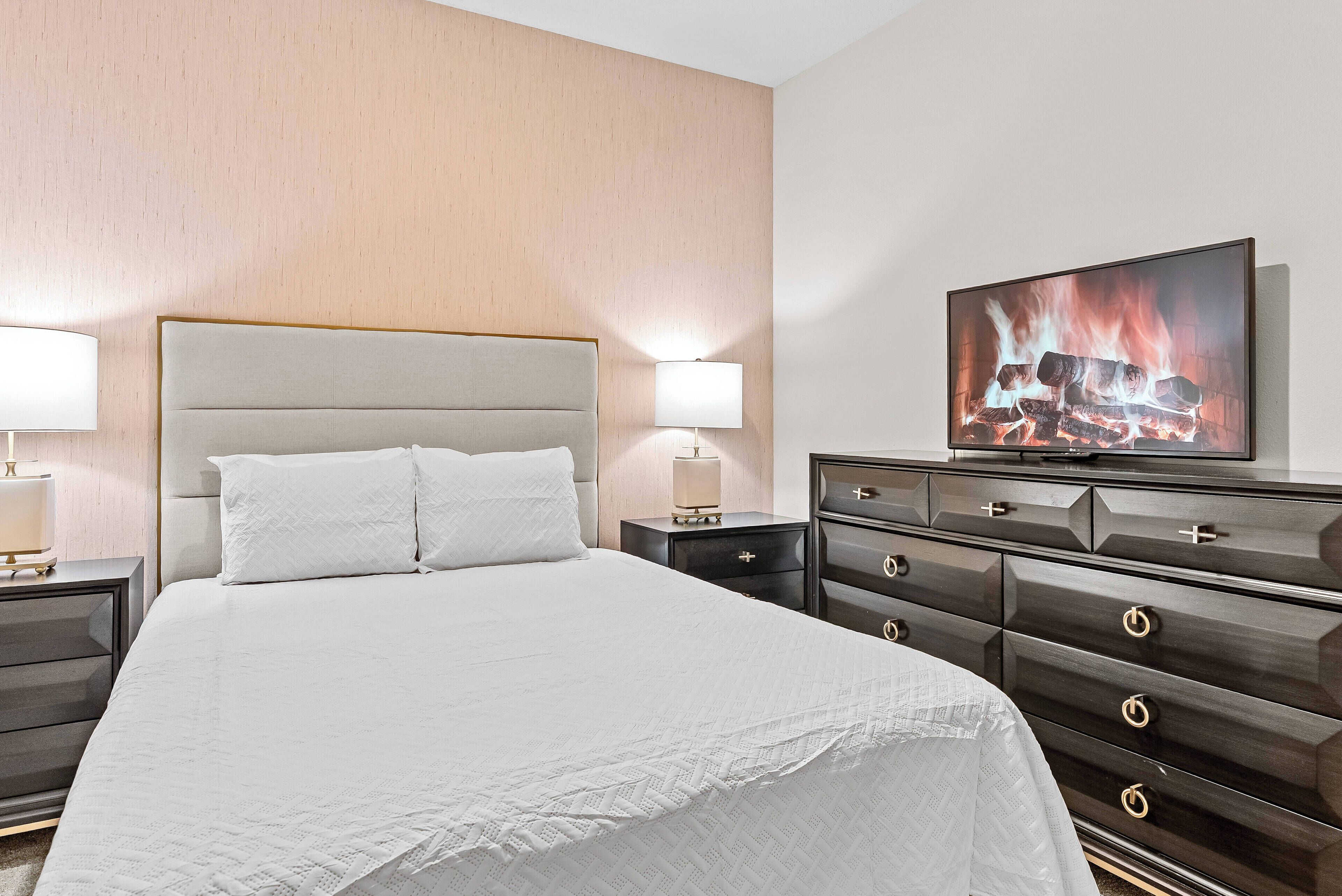 A hotel room with a large bed, two bedside tables with lamps, a dark wooden dresser, and a television displaying a fireplace.